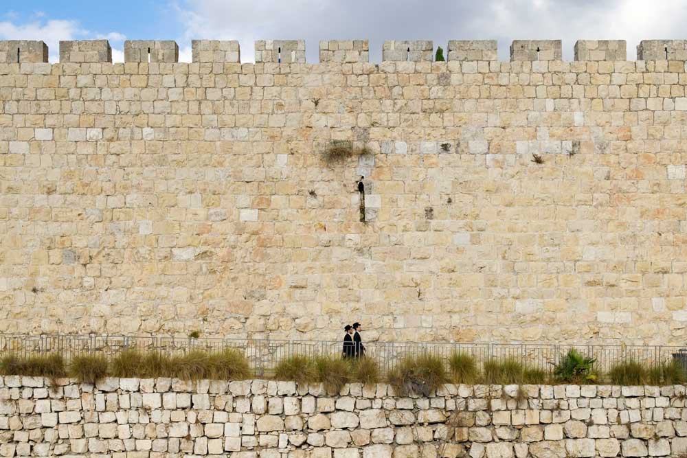 Dois homens com ternos e chapéus pretos, caminham ao lado de muro de pedras em Israel