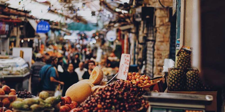 Frutas dispostas para venda, em barraca de feira. Ao fundo, embaçado, há pessoas andando pelo Mahane Yehuda Market, mercado em Jerusalém que é um dos passeios em israel