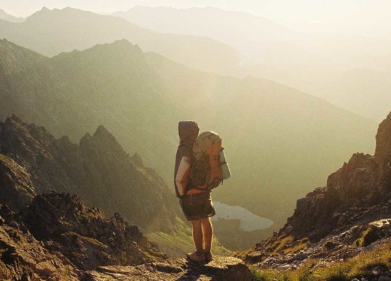 Viajante carrega mochila nas costas e observa, em cima de uma pedra, montanhas, durante o dia