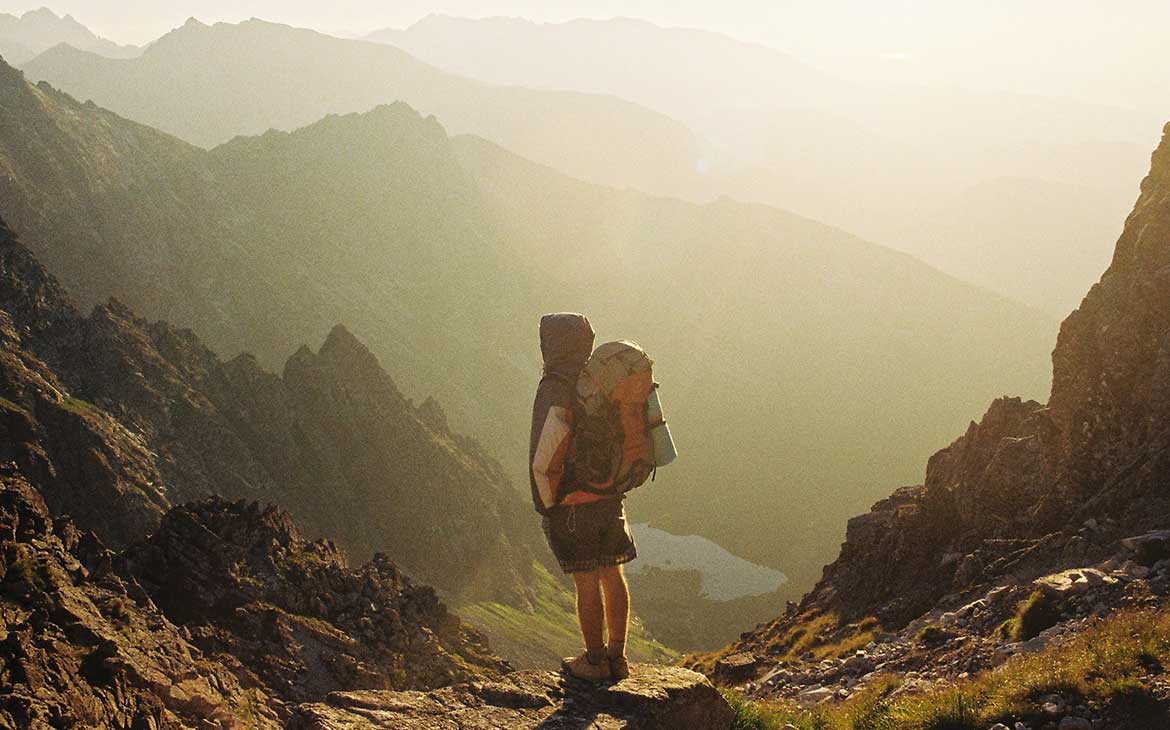 Viajante carrega mochila nas costas e observa, em cima de uma pedra, montanhas, durante o dia