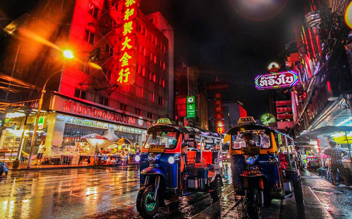Pessoas em tuk-tuks em estrada molhada, durante a noite, onde há estabelecimentos com fachadas de letreiros coloridos iluminados