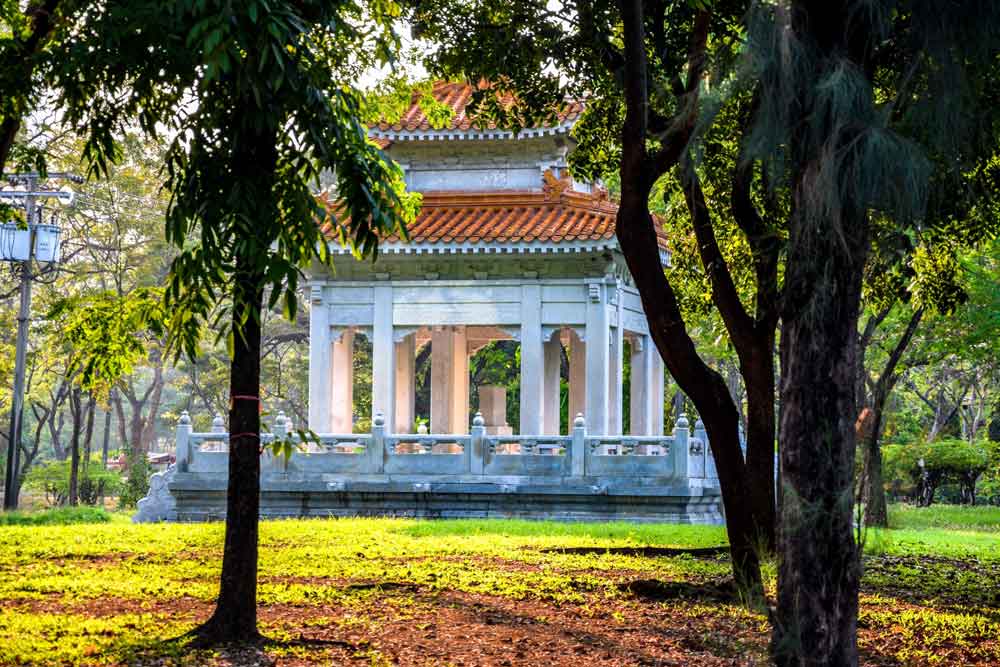 Construção com colunas brancas e com telhado marrom em meio ao gramado e árvores do Parque Lumpini, localizado em Bangkok