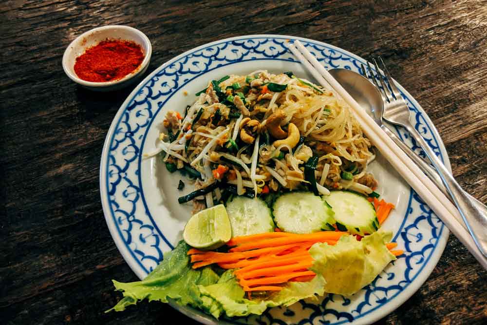 Pepino fatiado, cenoura, salada, macarrão e temperos em prato floral branco e azul ao lado de garfo, colher e hashis, em mesa de madeira de um restaurante em Bangkok