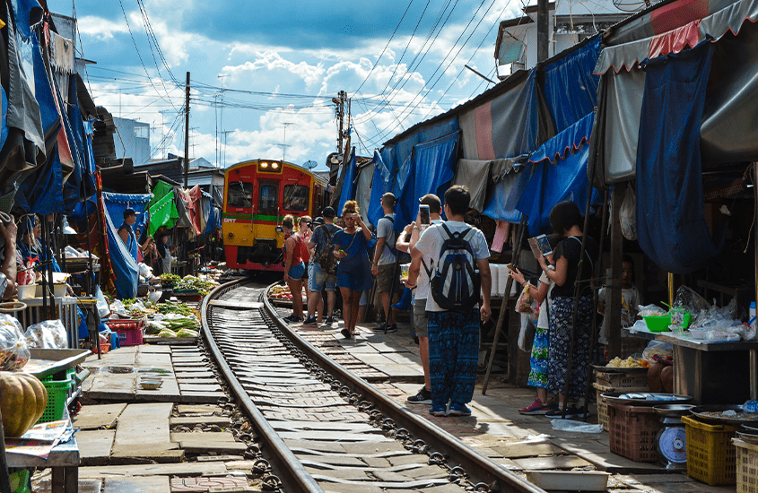 durante o dia, pessoas observam trem passando em trilhos, cercado por barraquinhas de mercado de trem bangkok