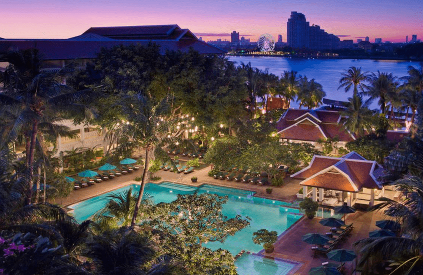 durante entardecer, vista aérea de árvores ao redor de piscina em hotel em bangkok e, ao fundo, silhueta de prédios em frente ao mar