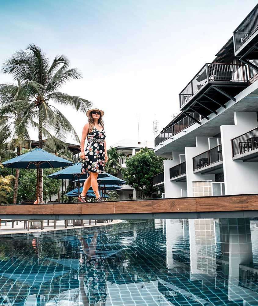 durante o dia, bárbara rocha, com chapéu branco, óculos de sol, vestido florido e sandália preta, caminha em frente a piscina de hotel em bangkok