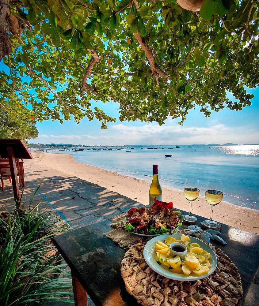 Em um dia de sol. restaurante onde comer em Búzios com praia na frente, barcos e árvores ao redor