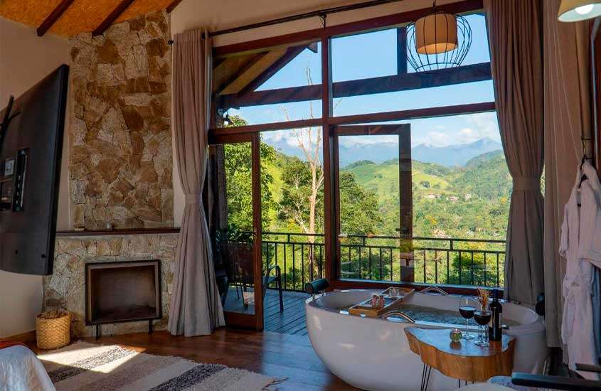 Interior de quarto com tapete, banheira, mesa, lareira de pedra, taças de vinho, roupões de toalha, TV e janelas grandes acortinadas com vista das montanhas