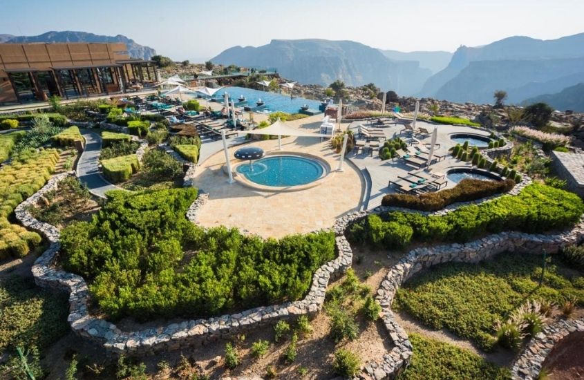 vista aérea de lazer de anantara al jabal al akhdar resort, onde há arvores, piscinas, espreguiçadeiras e montanhas ao fundo, durante o dia em omã