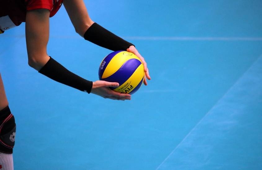 em quadro de vôlei, jogadora segura com as duas mãos uma bola azul e amarela