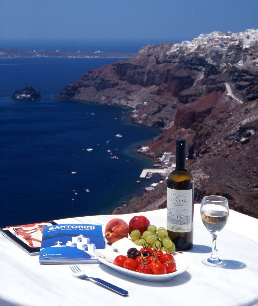 frutas e vinhos em cima de mesa branca com vista para o mar da grécia