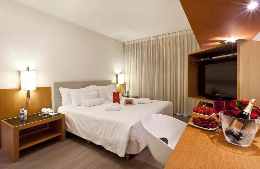 espumante e frutas em cima de mesa de quarto com cama de casal e televisão em h2 platinum lourdes, um dos melhores hotéis em belo horizonte
