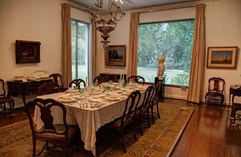 sala com mesa e cadeiras de madeira, quadros de arte e janela com vista para árvores em museus da chácara do céu