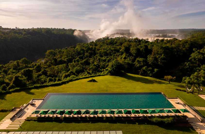 Em um dia de sol, área de lazer de hotel em foz do iguaçu perto das cataratas com piscina, guarda-sóis, espreguiçadeiras, árvores e cataratas ao redor