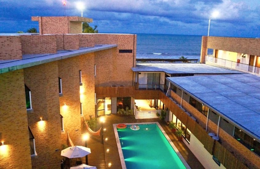 vista aérea de piscina em área de lazer de bessa beach hotel e mar ao fundo, durante anoitecer em joão pessoa