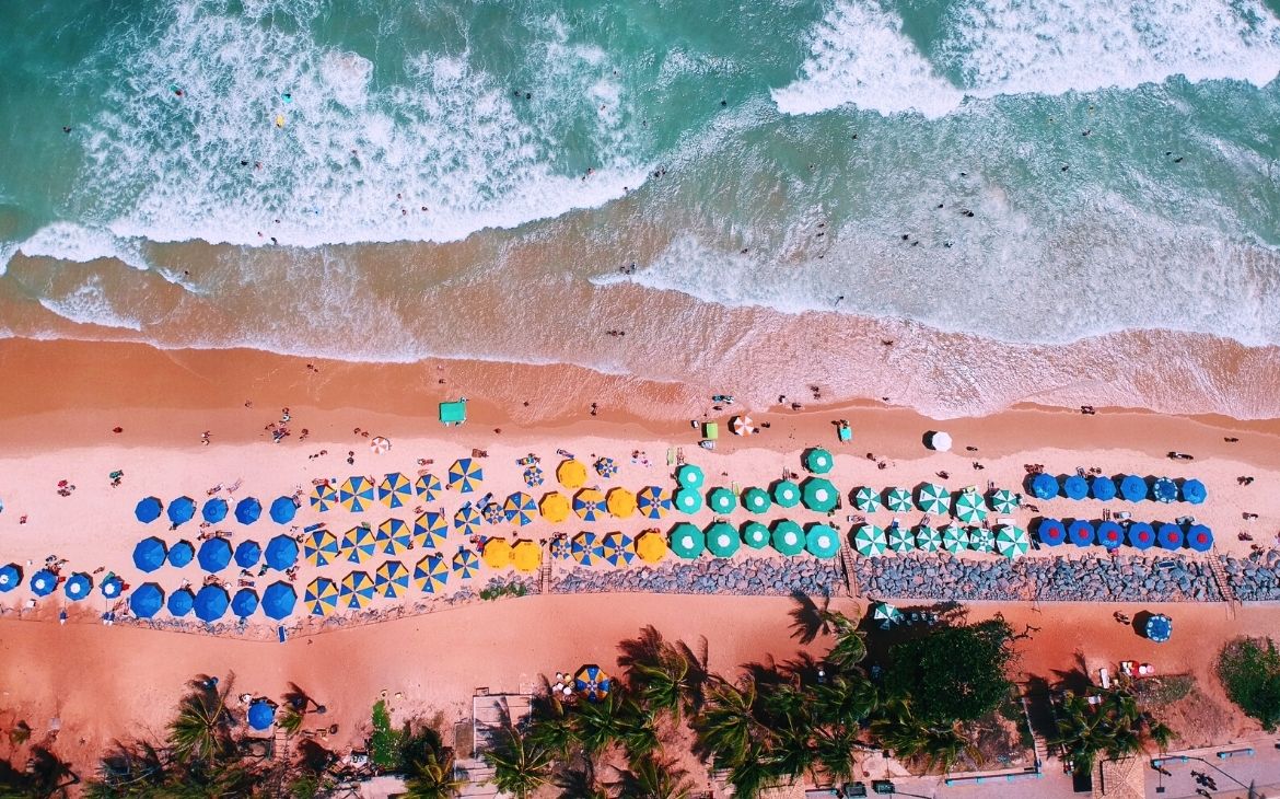 vista aérea de coqueiros e guarda-sóis coloridos em areia, e mar ao fundo em praia do nordeste
