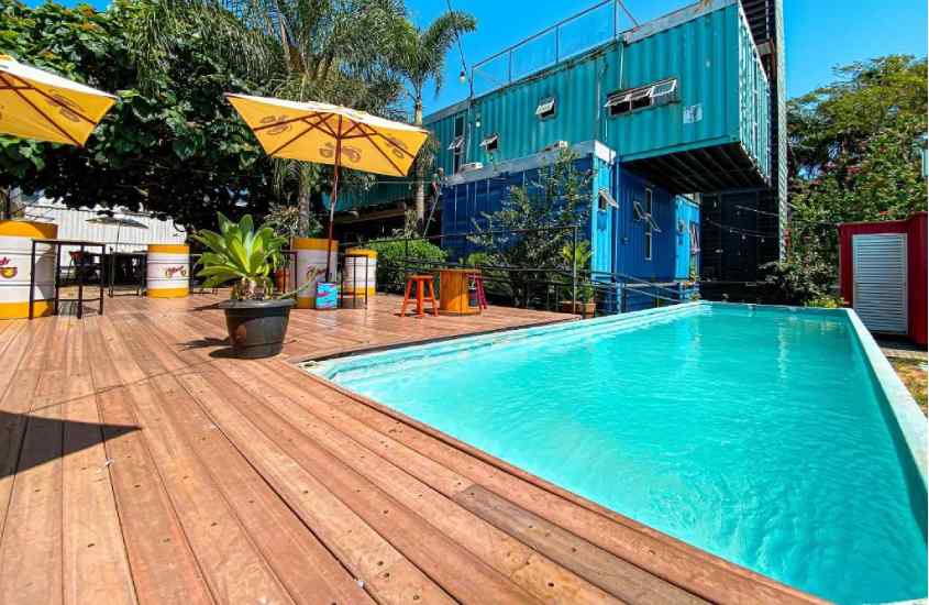Em um dia ensolarado, área de lazer de um hostel com piscina, deck de madeira, guarda-sól, plantas decorativas, mesas e cadeiras