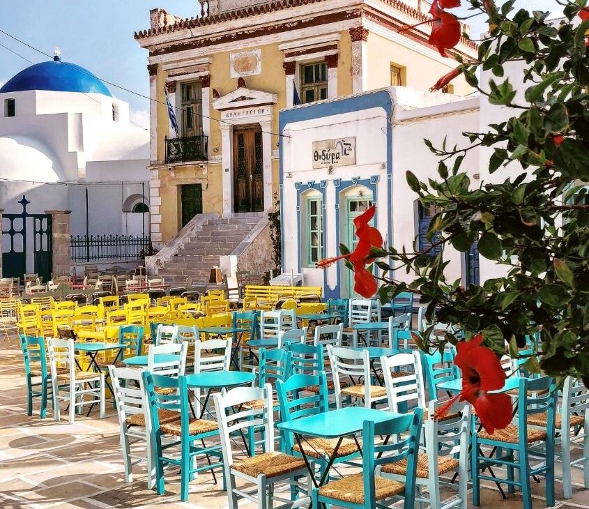 mesas e cadeiras coloridas em rua cheia de restaurantes, durante o dia, em santorini