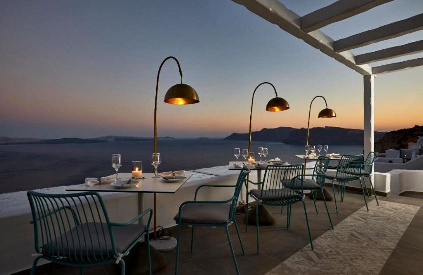 cadeiras e mesas onde há taças e pratos, durante entardecer, em restaurante de filotera suites um dos melhores hotéis em santorini para quem vai passar a lua de mel na grécia