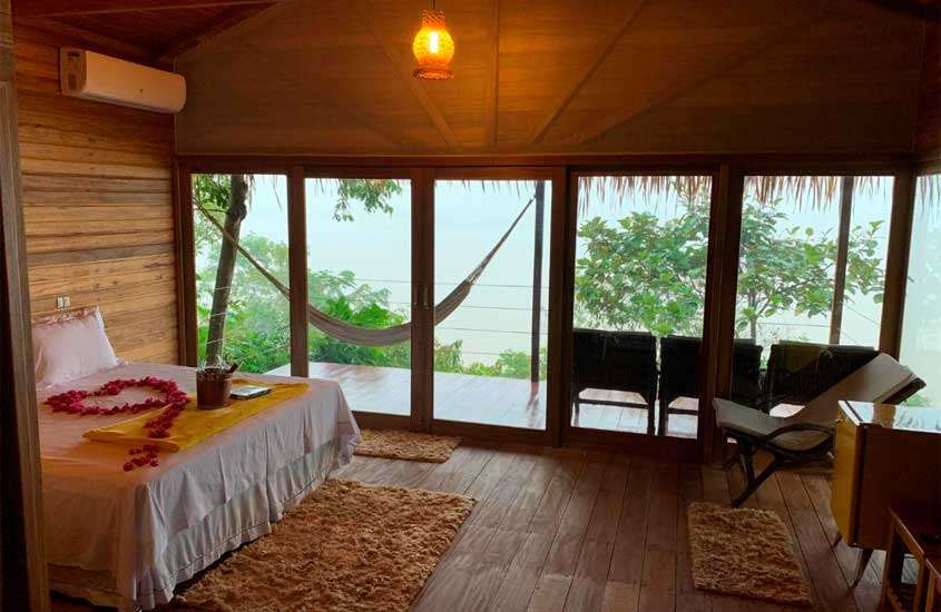 Quarto de um dos hotéis de selva em manaus com cama de casal, tapetes, cadeira, frigobar, ar-condicionado, rede e cadeiras na varanda