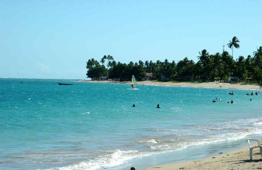 Em um dia ensolarado, praia com pessoas no mar, árvores, barco e pessoa praticando windsurf