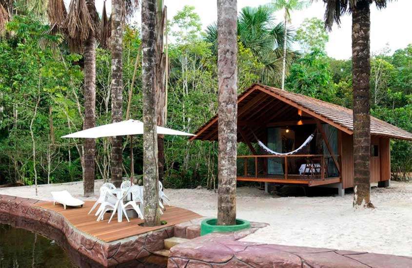 Em um dia nublado, área de lazer de um dos hotéis de selva no amazonas com mesas, cadeiras, espreguiçadeira, guarda-sol, árvores ao redor e cabana de madeira com rede na varanda