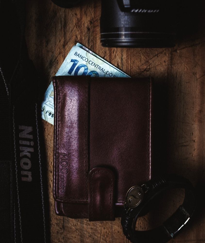 carteira, nota de 100 reais, camera nikon e pulseira de couro em cima de mesa de madeira