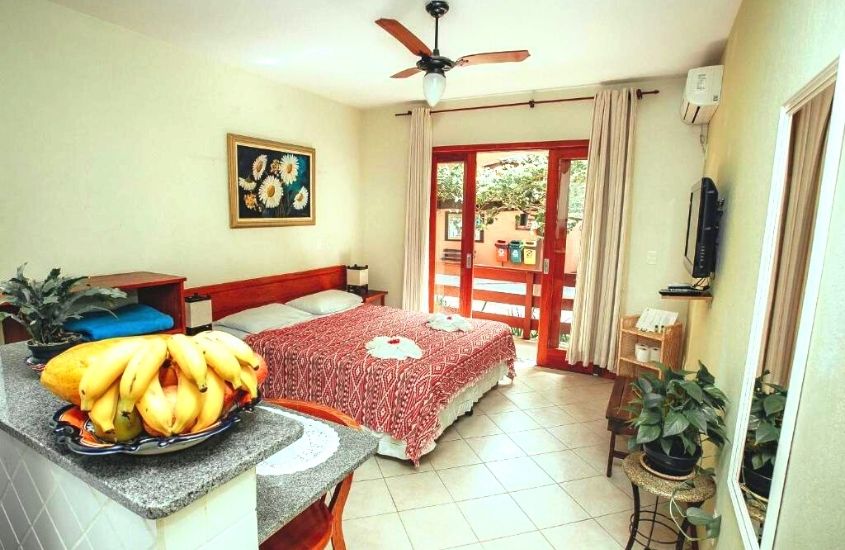 mesa com fruteira, cadeira, cama de casal, planta, televisão e ventilador de teto em suíte com varanda de Pousada Colinas da Lagoa