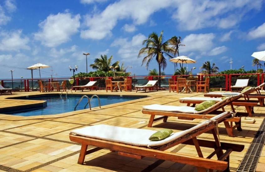 espreguiçadeiras, mesas e cadeiras ao redor de piscina, durante dia ensolarado em área de lazer do Bahiamar Hotel