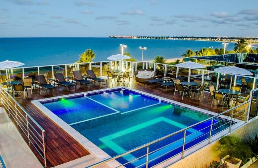 piscina, cadeiras, mesas e espreguiçadeiras em área de lazer com vista para o mar, durante anoitecer, em Laguna Praia Hotel