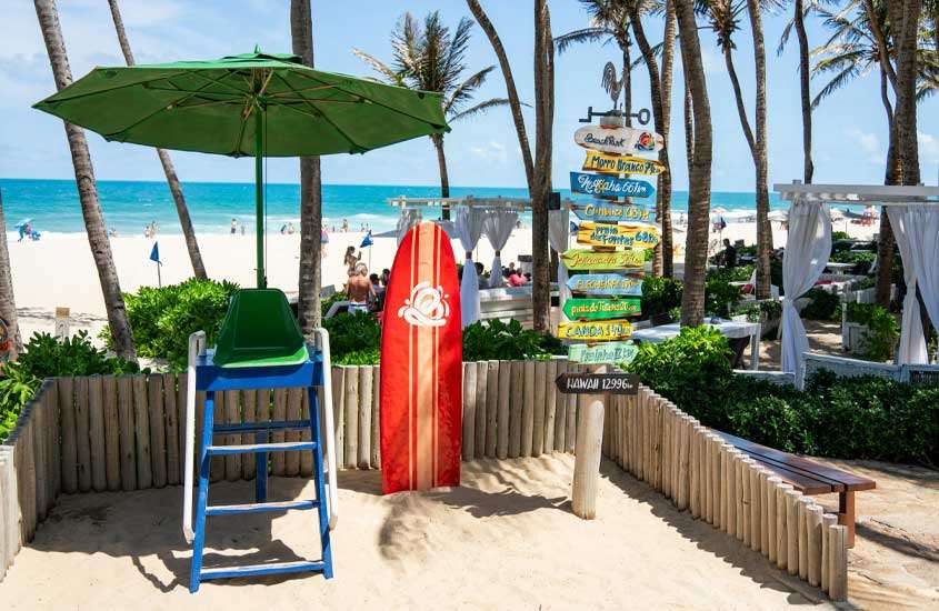 Em um dia de sol, entrada da praia com placas de madeira, tenda, árvores, prancha decorativa, cadeira de salva-vidas e mar no fundo