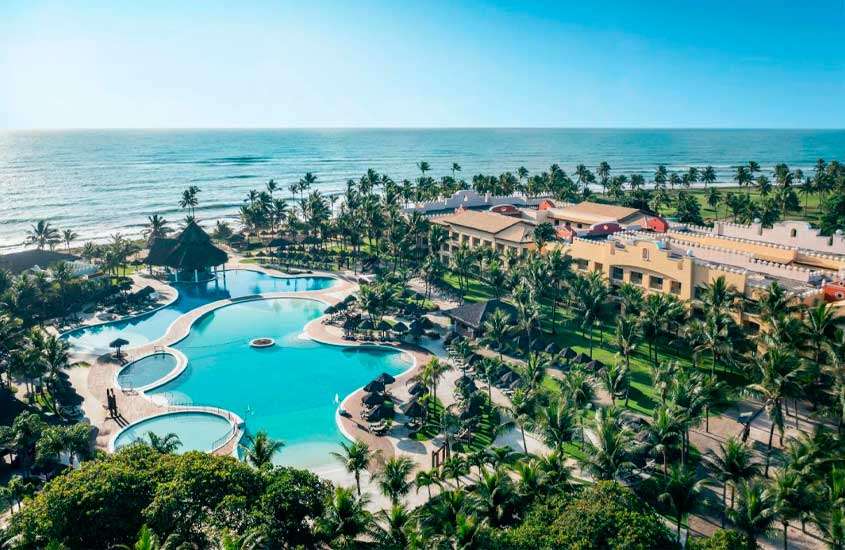 Em um dia ensolarado, vista aérea de hotel com praia na frente, piscinas, árvores e tendas ao redor