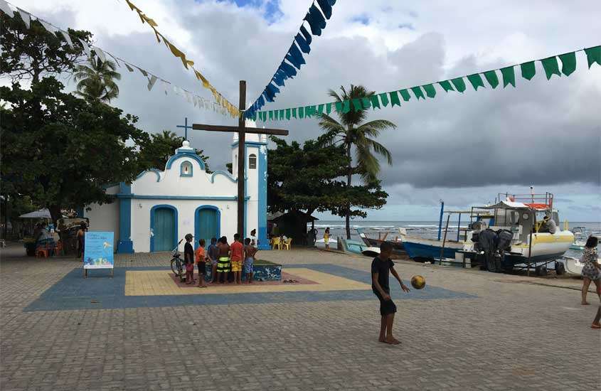 Em um dia nublado, praça em frente a igreja com bandeirinhas, pessoas e barcos aportados
