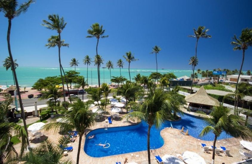 vista aérea de piscina rodeada de coqueiros, em frente ao mar, em área de lazer de Maceió Atlantic Suites