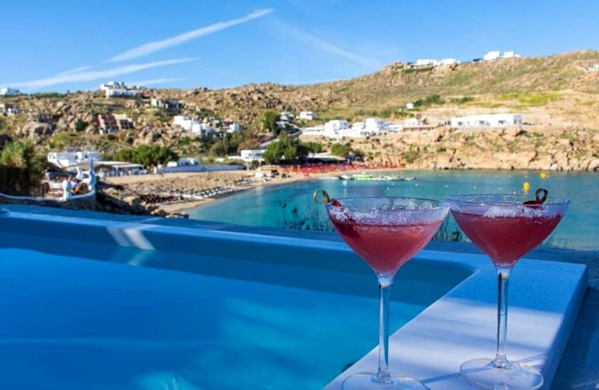 taças de vidro com bebida vermelha, em cima de beira de piscina com vista para o mar, durante o dia em suíte de Nimbus My Aktis um dos hotéis em mykonos