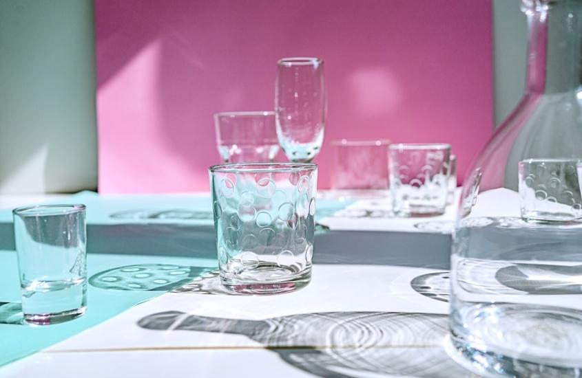 copos, taças e garrafas de vidro em cima de mesa branca, ao fundo parede rosa