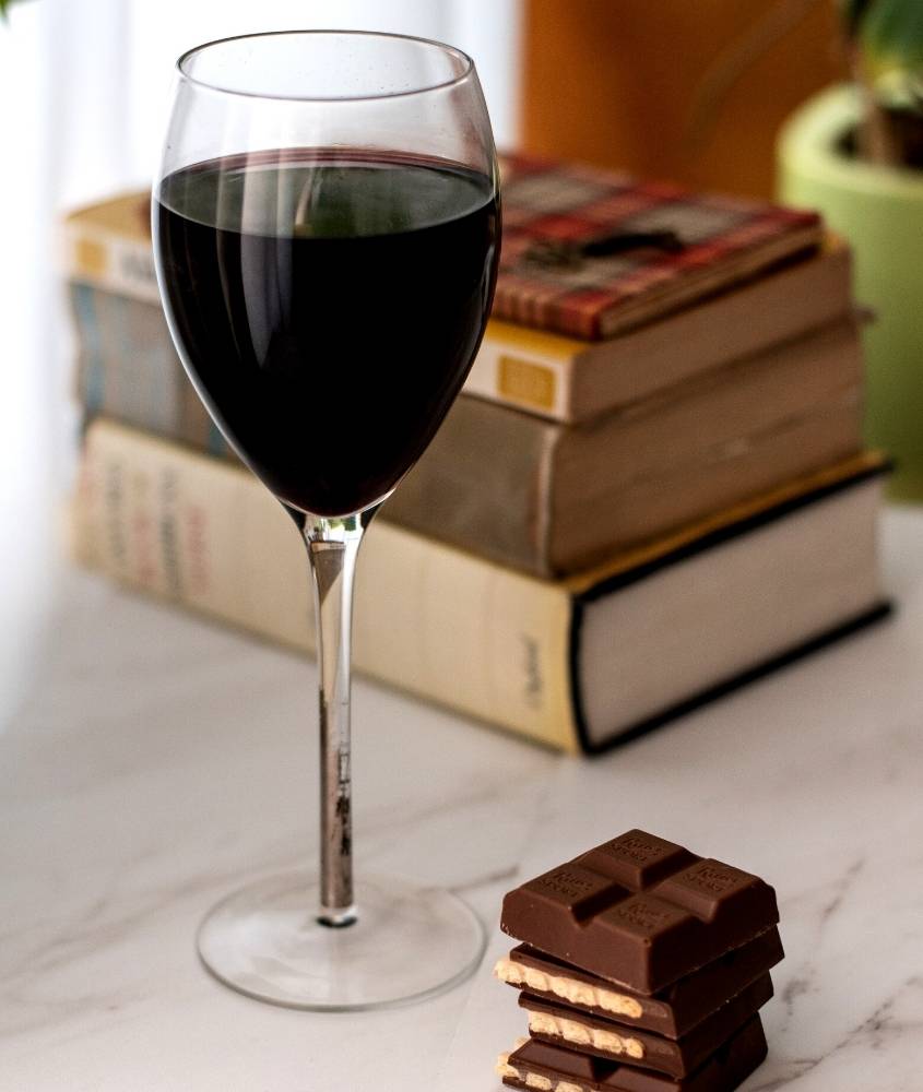 taça de vidro com vinho, pedaços de chocolate e livros em cima de mesa