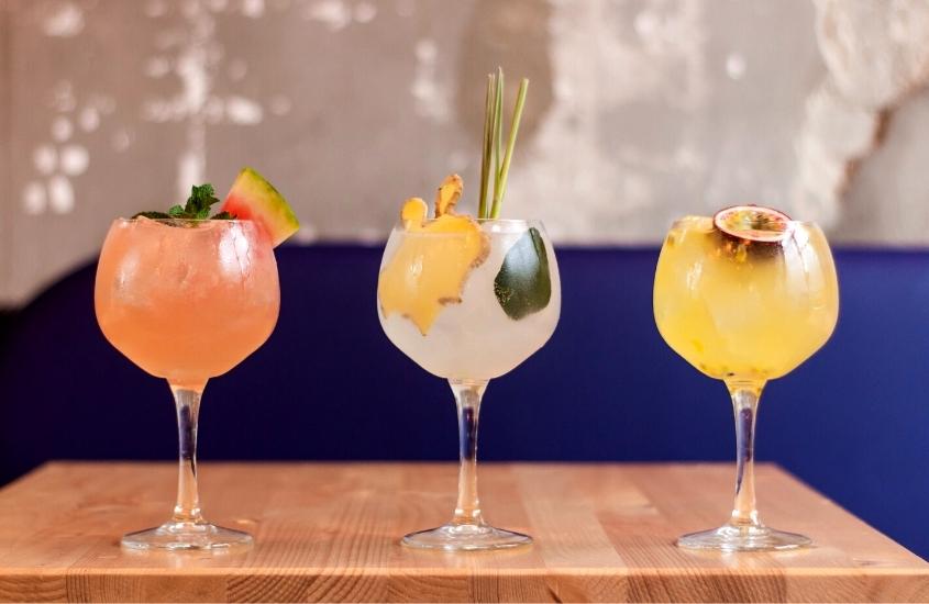 três taças de vidro com drinks e frutas em cima de mesa de madeira em bar, uma opção para quem busca o que fazer em garibaldi a noite