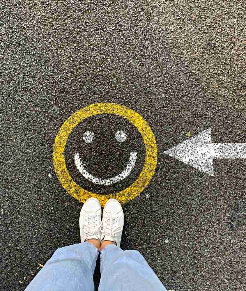 pessoa com calça jeans e tênis branco, pisando em asfaltando onde há pintado desenho de emoji amarelo e branco sorrindo