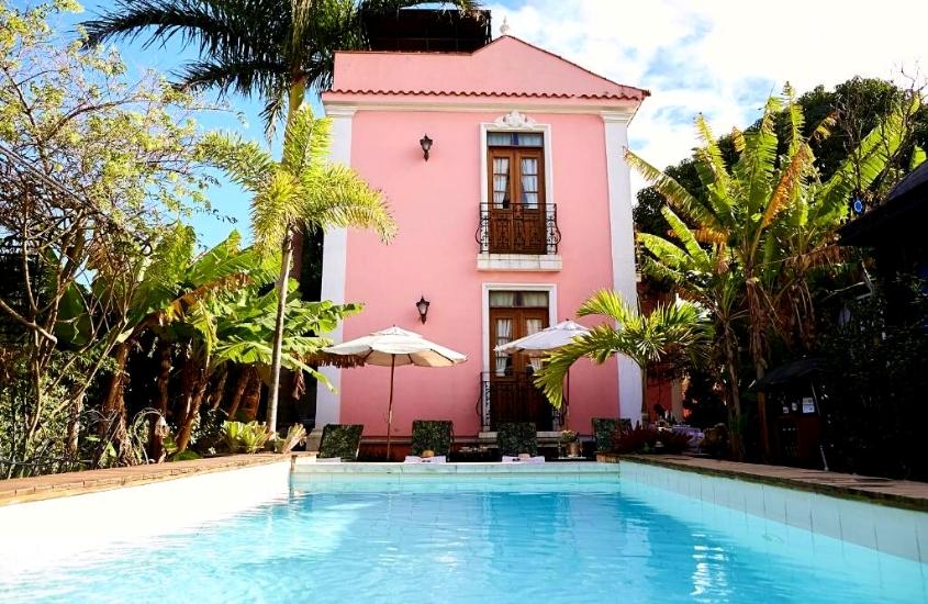 durante o dia, piscina cercada de árvores em frente a casarão rosa de dois andares onde funciona a Hotel Boutique Quinta das Videiras