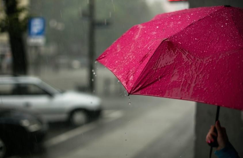 pessoa segura guarda-chuva rosa, ao fundo embaçado, carros passam em rua