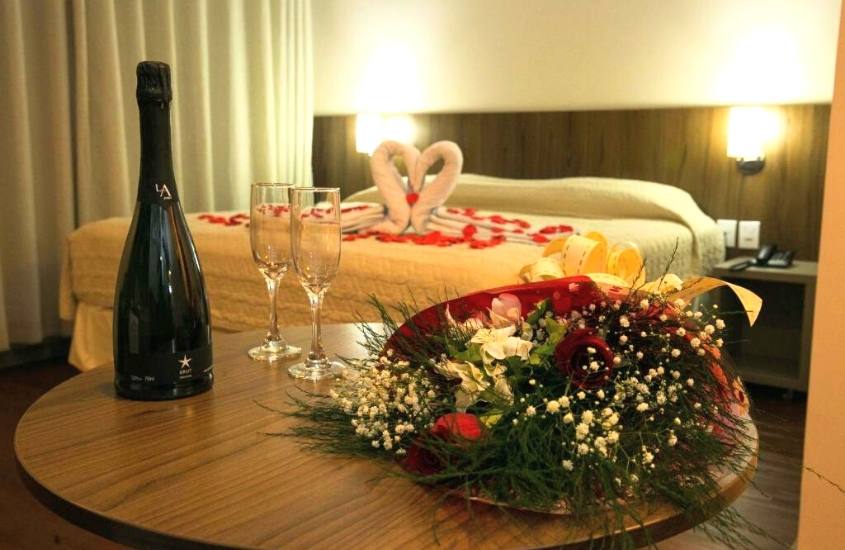 garrafa de champanhe, duas taças de vidro e buquê de flores em cima de mesa de madeira redonda e cama de casal ao fundo em suíte de Hotel Dom Rafael Premium