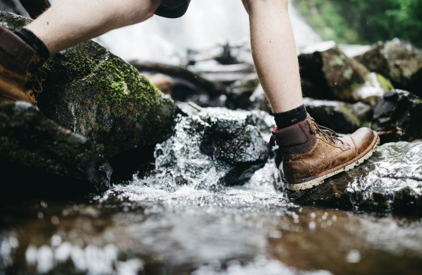 pernas de joelho para baixo de pessoa de bota marrom atravessando rio durante caminhada