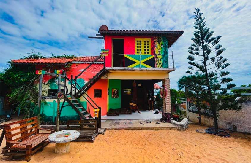 Em um dia de sol com nuvens, parte de trás de hostel com pintura colorida, bancos, mesas, árvores e parte de areia