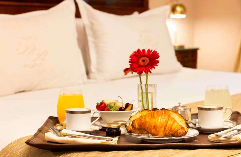 Cama de casal com bandeja de café da manhã com croissants, sucos, cafés, frutas e flores