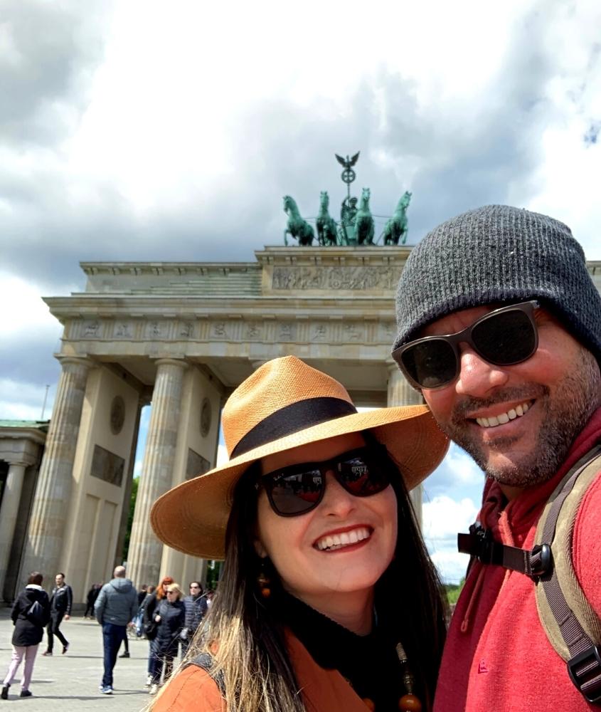 bárbara rocha, com chapéu e óculos de sol, e vagner alcantelado com touca cinza e óculos de sol, sorriem em frente a portão de brandemburgo, durante o dia