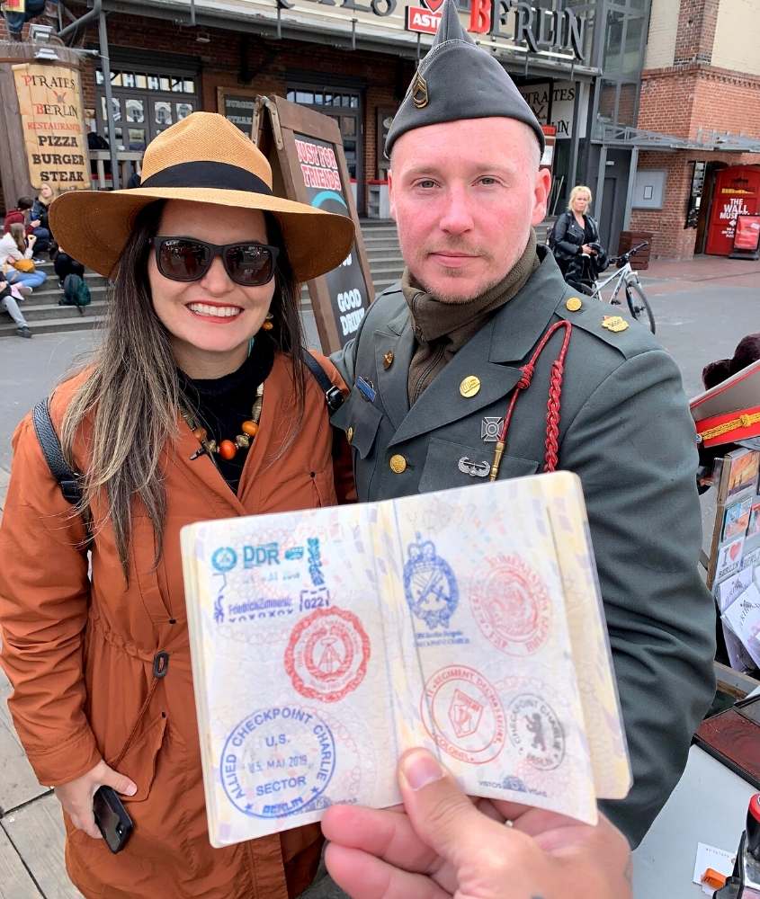 bárbara rocha, de óculos de sol, chapéu e casaco laranja, sorri ao lado de militar alemão uniformizado em checkpoint charlie, durante o dia
