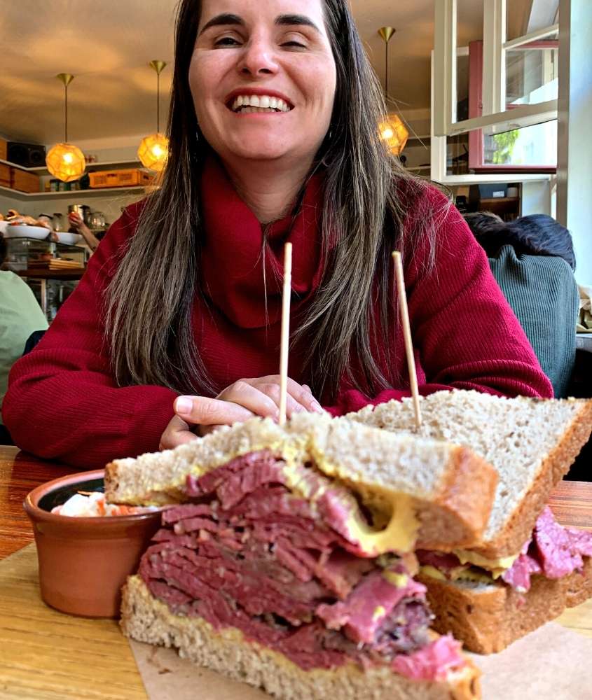 bárbara rocha, com cacaso vermelho, sorri sentada em frente a mesa onde há em cima sanduíche de pastrami com queijo do restaurante mogg