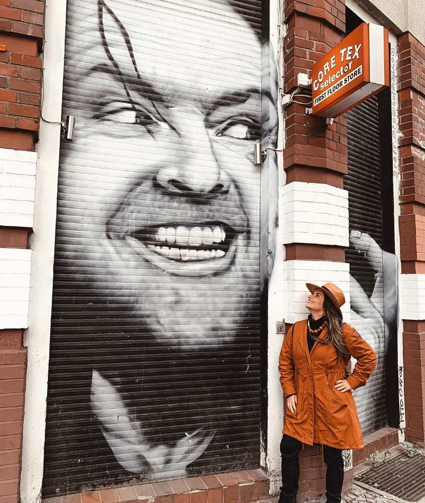 bárbara rocha, com chapéu, casaco laranja e calça jeans, sorri em frente a mural em rua oranienburger