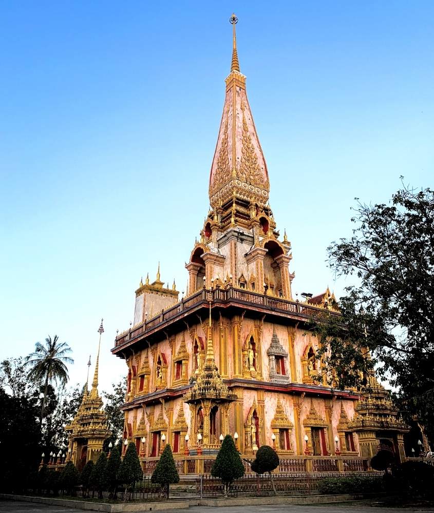 durante o dia, árvores em frente a construção com detalhes dourados onde funciona o templo Wat Chalong